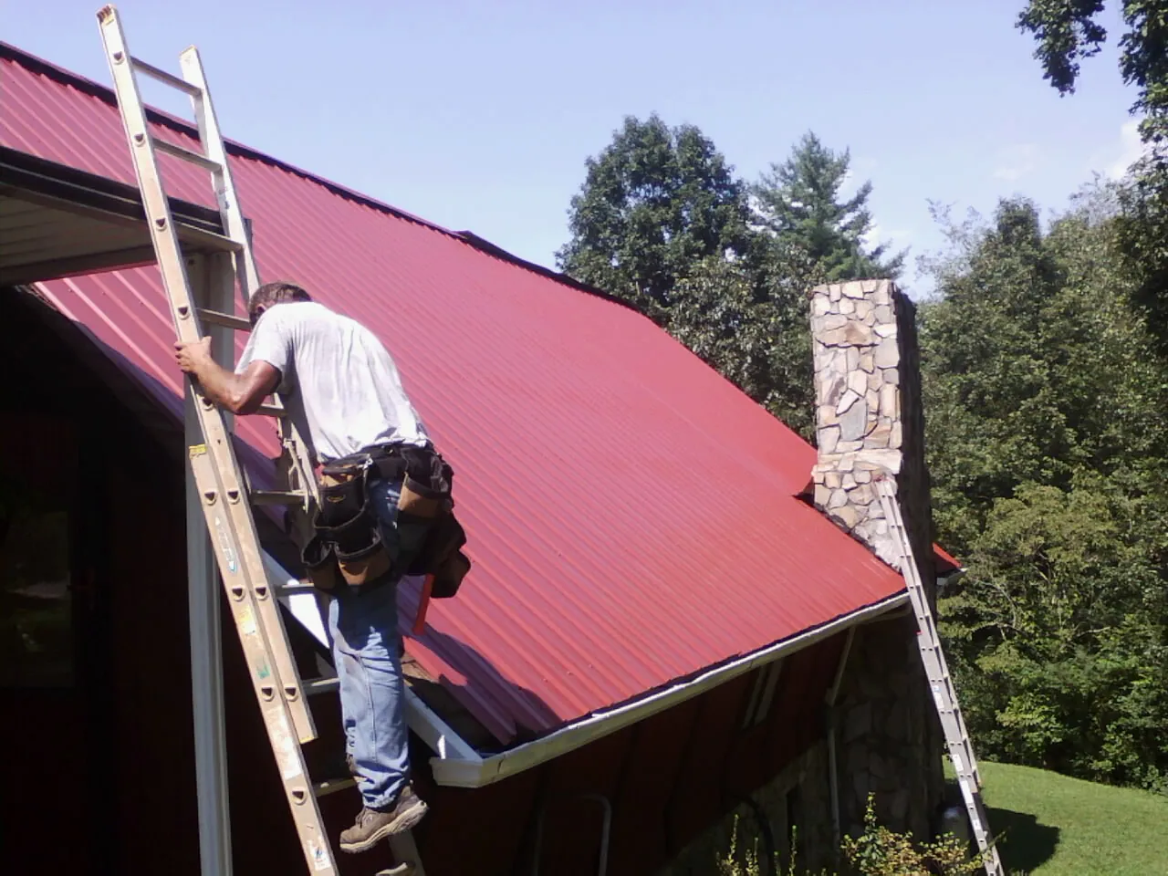 Roofer on a ladder beside a red metal roof and stone chimney — Soffit & Fascia Repair by Roofing Brothers in Brookside